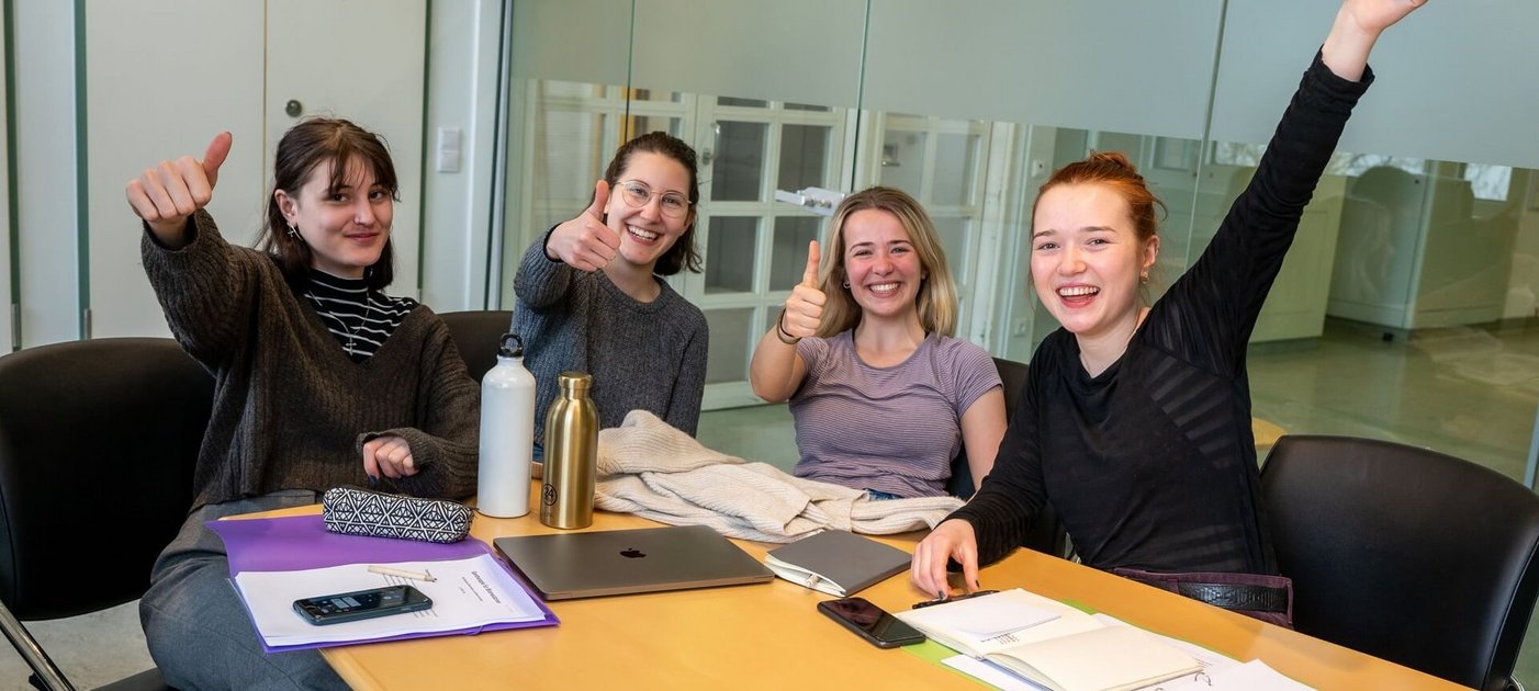 Studierende sitzen um einen Tisch in einem Lernraum. Am Tisch liegen Lernunterlagen. Die Studierenden freuen sich und jubeln der Kamera zu.