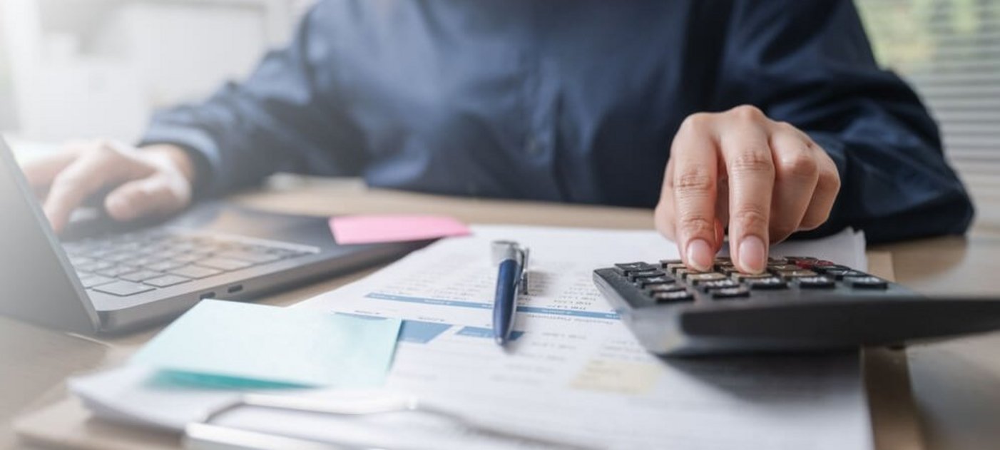 Jemand sitzt mit Unterlagen und einem Taschenrechner vor einem Laptop