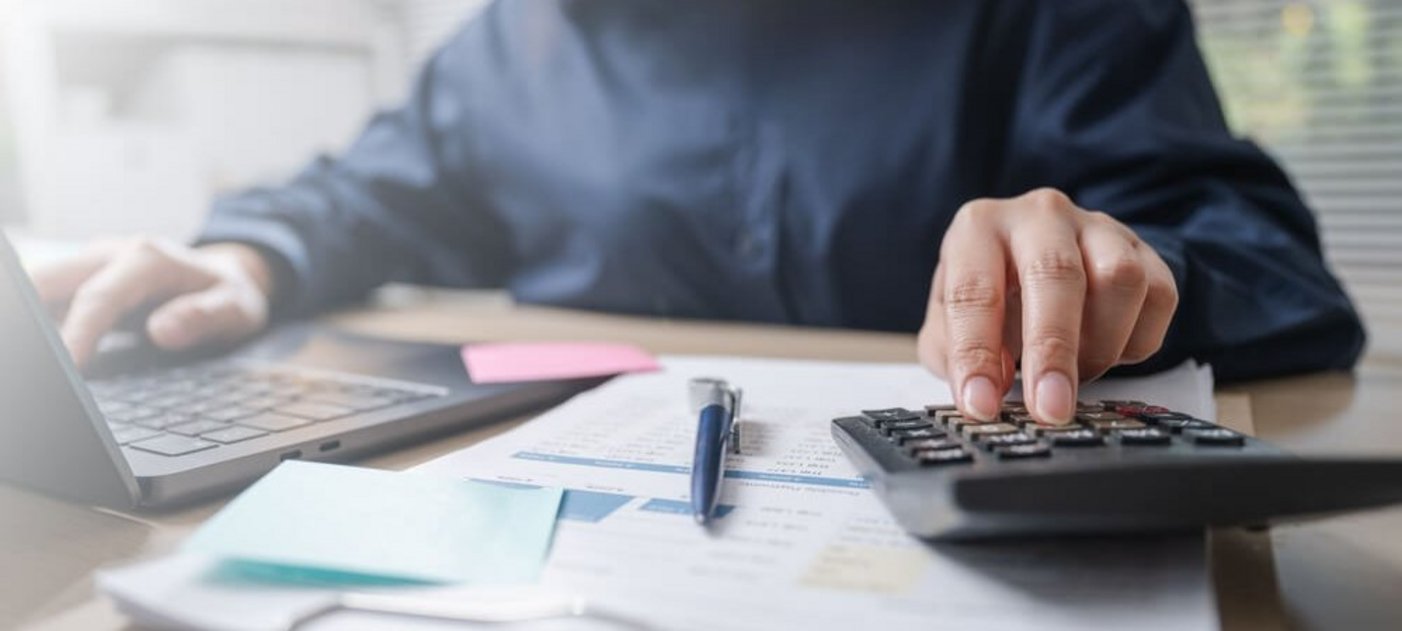 Jemand sitzt mit Unterlagen und einem Taschenrechner vor einem Laptop
