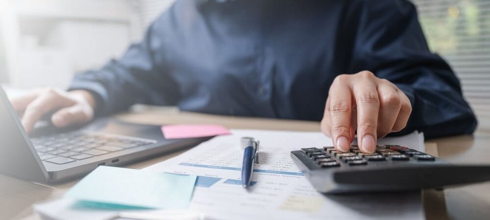 Jemand sitzt mit Unterlagen und einem Taschenrechner vor einem Laptop