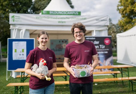 Viktoria Weissenb&ouml;ck und Patrick Birkl vor dem Vetmeduni-Zelt am ernte.dank.festival 2026 mit einem H&uuml;hner-Stofftier und einer Box mit Einstreu. 